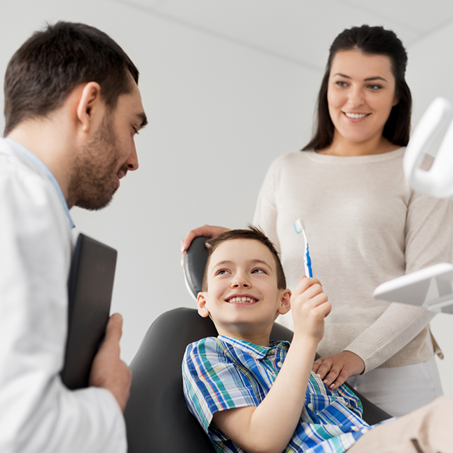 Boy with toothbrush sitting in dental chair with mother and dentist next to him